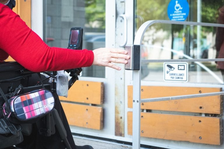 Gros plan sur la main d’une femme en fauteuil roulant appuyant sur un bouton-poussoir pour ouvrir une porte.