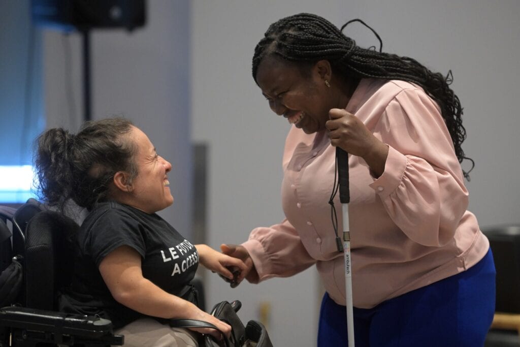 Deux femmes se sourient en discutant. L’une utilise une canne blanche et l’autre un fauteuil roulant électrique.